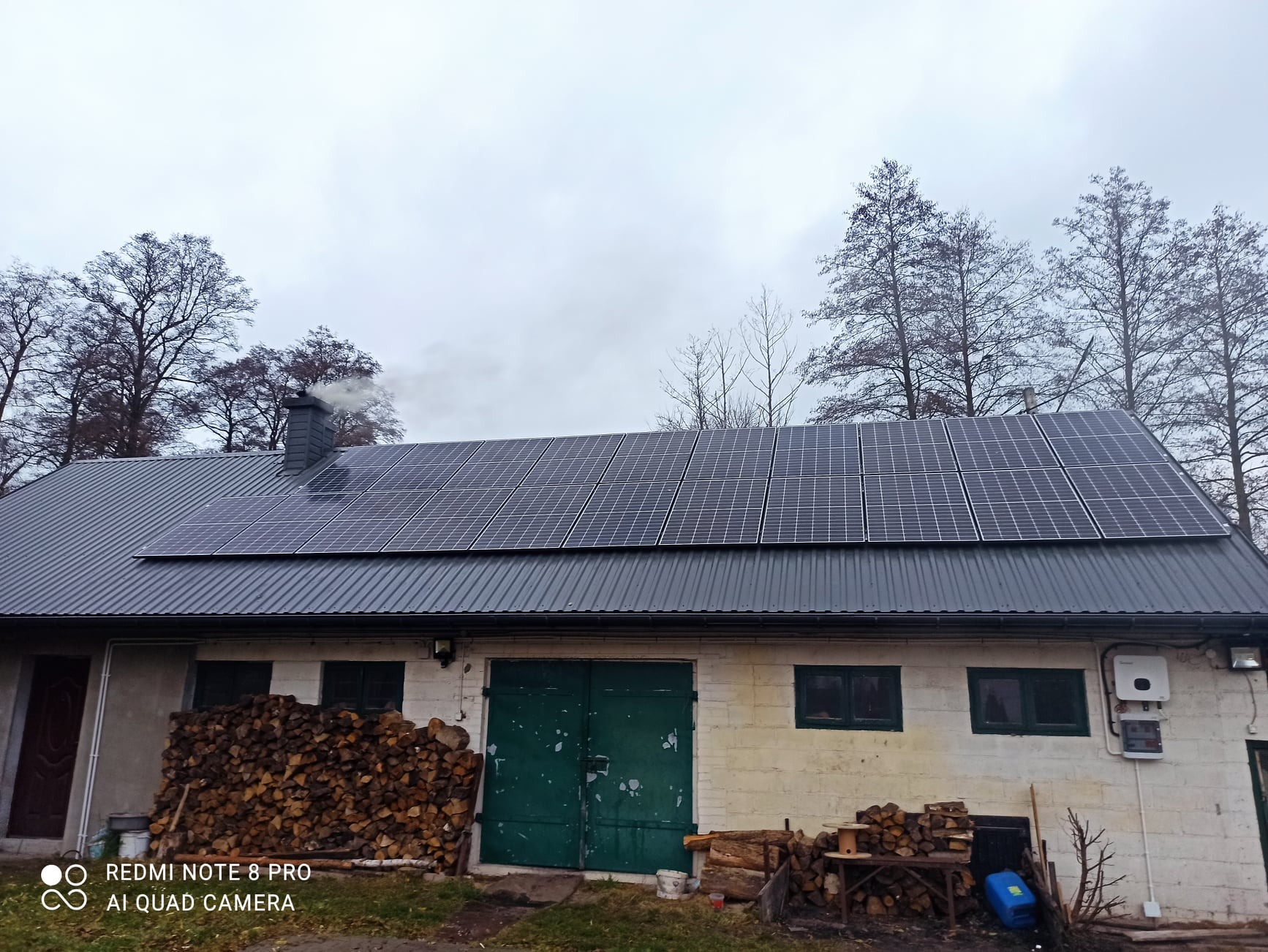 Instalacja paneli słonecznych na dachu budynku gospodarczego z kominem, obok sterta drewna, pochmurne niebo.