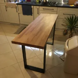 Blat drewniany na metalowych nogach w jasnej kuchni z białymi szafkami i płytkami podłogowymi, widoczny fragment zlewu i innych akcesori&oacute;w kuchennych