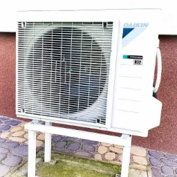 Biała zewnętrzna jednostka klimatyzacji Daikin R32, zamontowana na metalowym stelażu, widoczna na tle ściany o fakturze baranka i kostki brukowej z mchem.