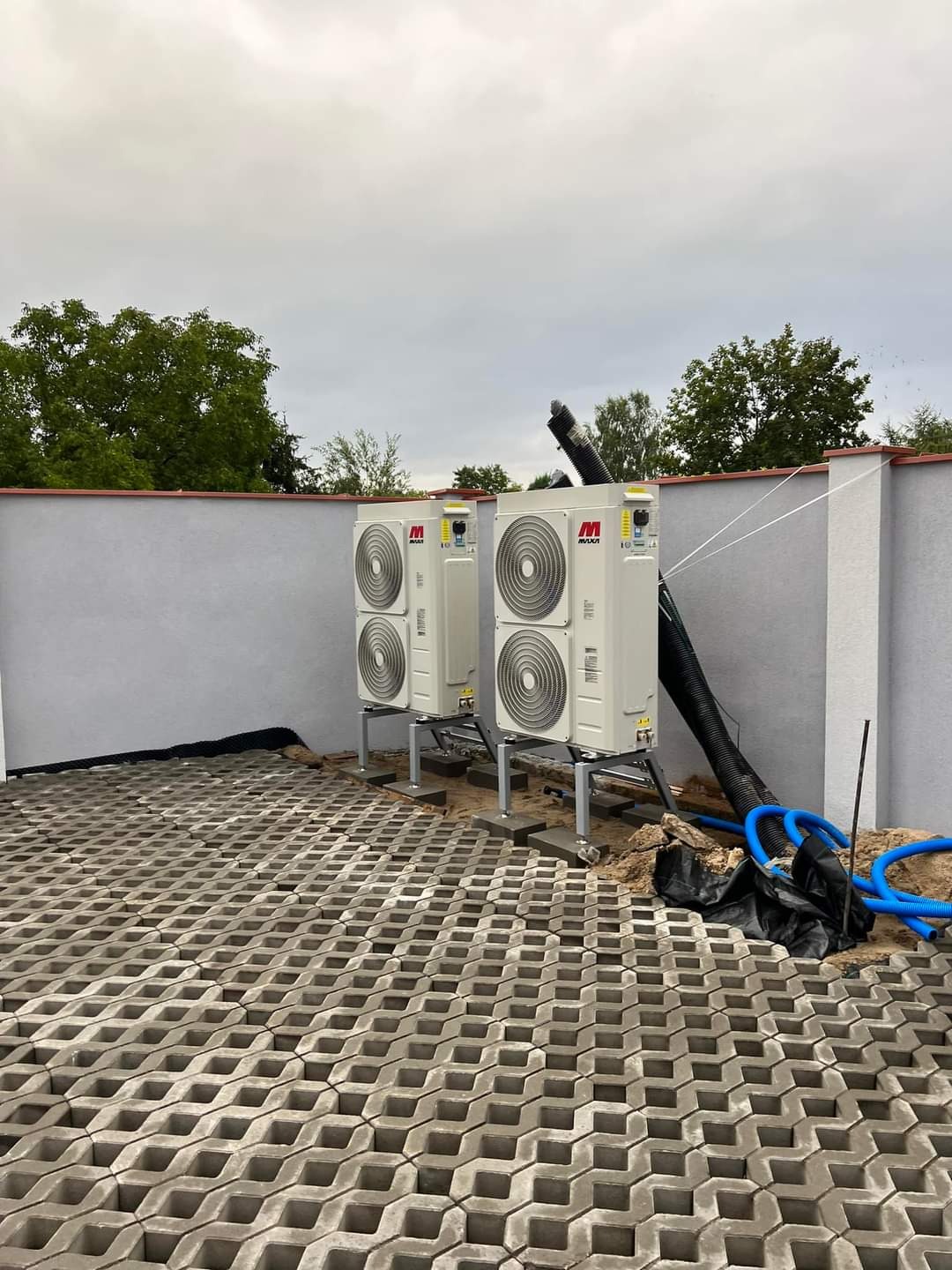 Dwie białe jednostki zewnętrzne z logo MAXA zamontowane na metalowych konstrukcjach na betonowej kostce ażurowej; widoczne niebieskie rury i czarna folia, szary mur w tle.