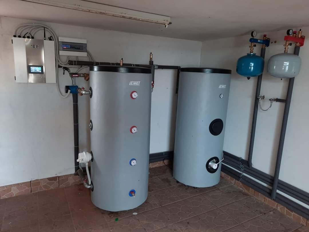 Dwa szare zbiorniki buforowe EMEI z manometrami w kotłowni, podłączone do instalacji z rur izolowanych szarą pianką, z widoczną skrzynką sterowniczą na ścianie.