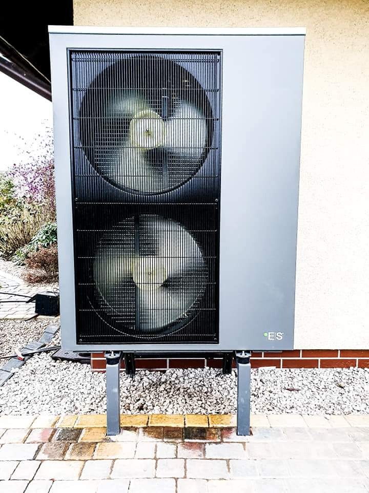 Szara pompa ciepła typu monoblok z dwoma wentylatorami, zamontowana na metalowych wspornikach na tle jasnej ściany budynku i kamiennego podłoża.