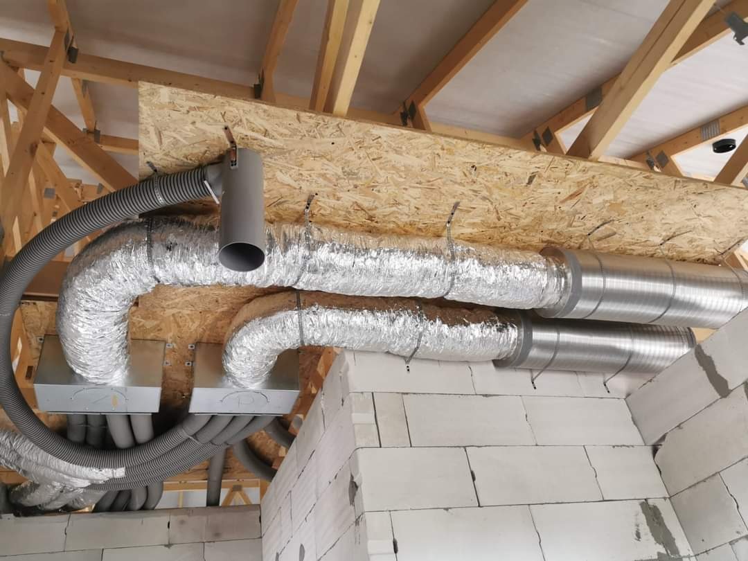 Szare, elastyczne przewody wentylacyjne podwieszone pod drewnianą konstrukcją dachu, widoczne elementy mocujące i izolacja termiczna na niektórych odcinkach.