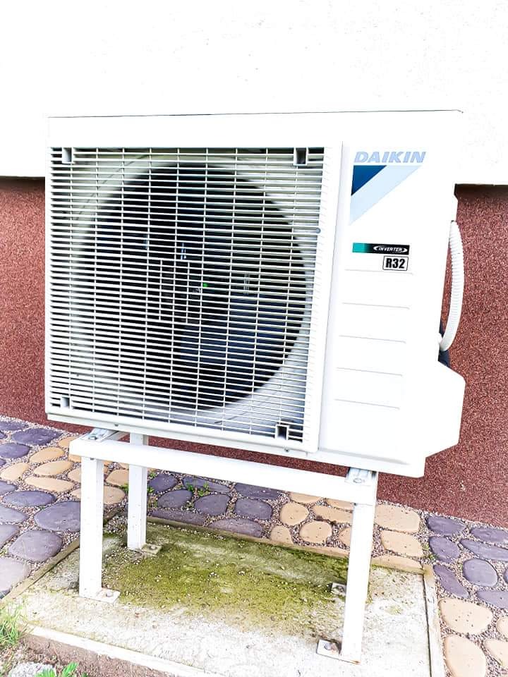 Biała zewnętrzna jednostka klimatyzacji Daikin R32, zamontowana na metalowym stelażu, widoczna na tle ściany o fakturze baranka i kostki brukowej z mchem.