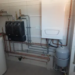 Kompletna instalacja grzewcza z kotłem, zasobnikiem i rozdzielaczem, widoczne rury miedziane i stalowe, zamontowane na ścianie z płytek.
