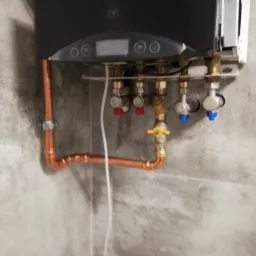 Zamontowany, nowoczesny piec gazowy z miedzianymi rurami i zaworami z czerwonymi i niebieskimi pokrętłami na tle ściany z szarego marmuru.