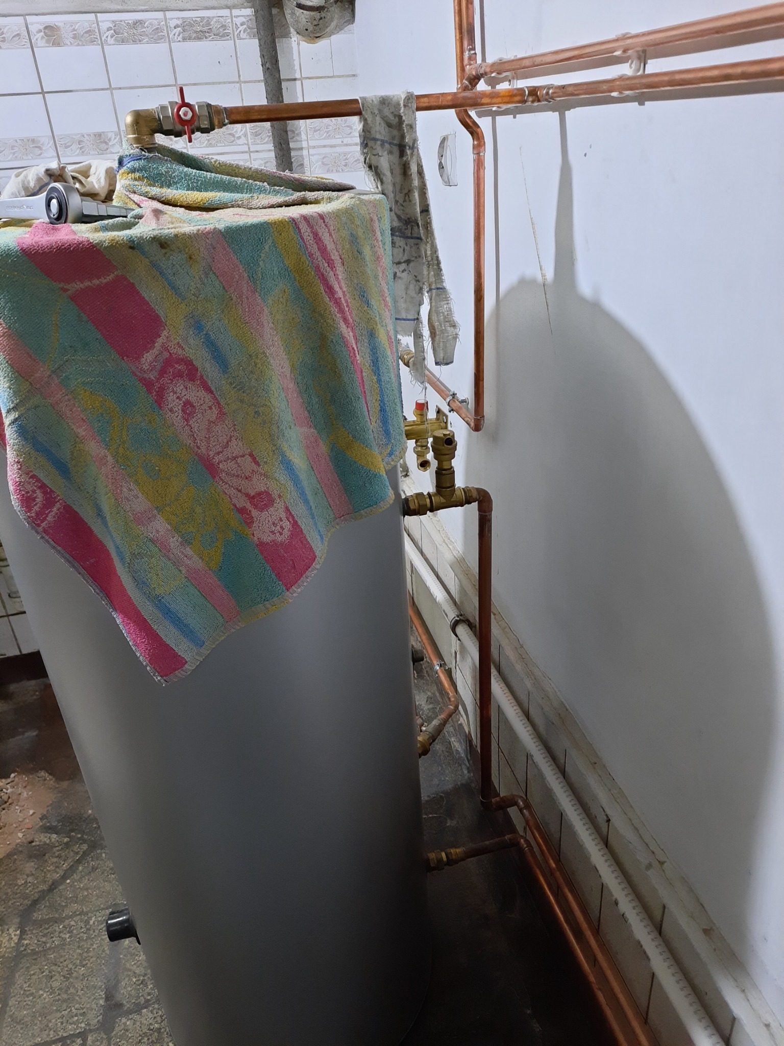 Szary zbiornik na wodę zakryty kolorowym ręcznikiem, z widocznymi miedzianymi rurami instalacji grzewczej i zaworami w tle, w pomieszczeniu z płytkami.