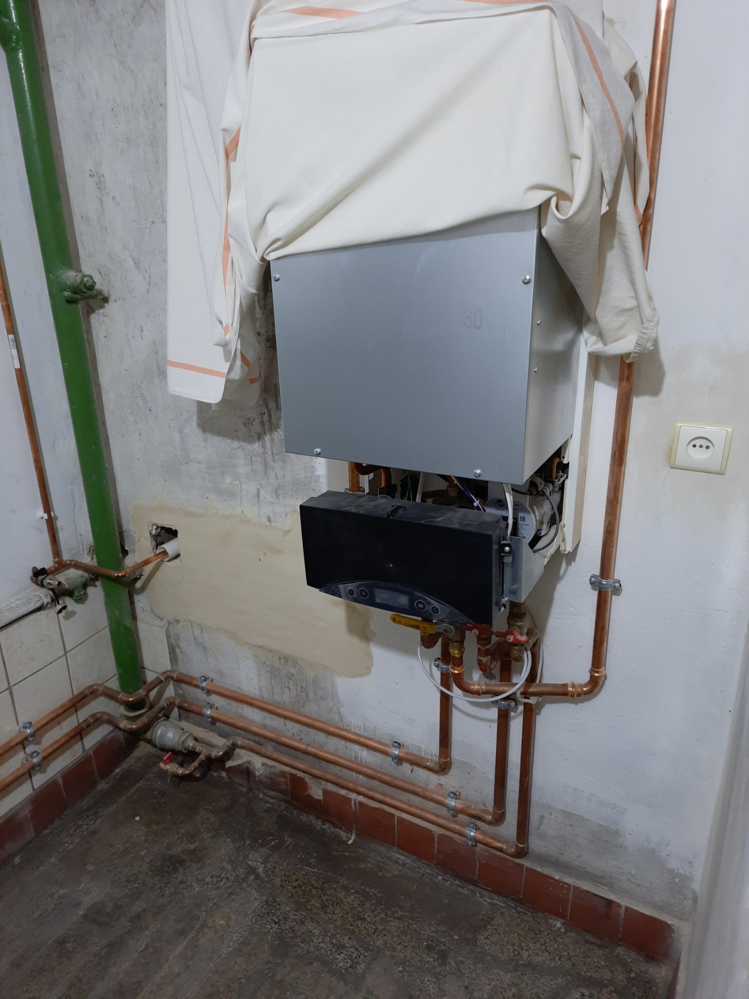 Nowy piec gazowy wiszący na ścianie, częściowo zakryty materiałem ochronnym, z widoczną instalacją hydrauliczną z miedzianych rur.