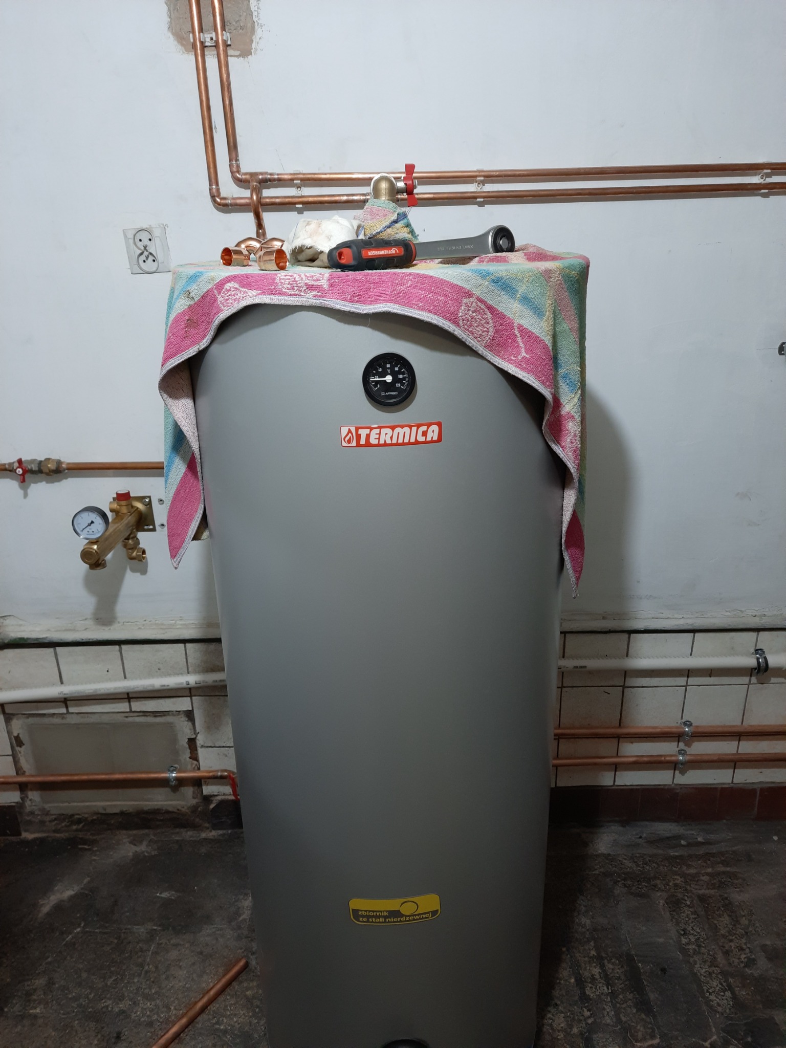 Szary zasobnik ciepłej wody użytkowej marki Termica z podłączonymi miedzianymi rurami, na górze leżą narzędzia i złączki zakryte ręcznikiem, widoczne manometry i zawory.