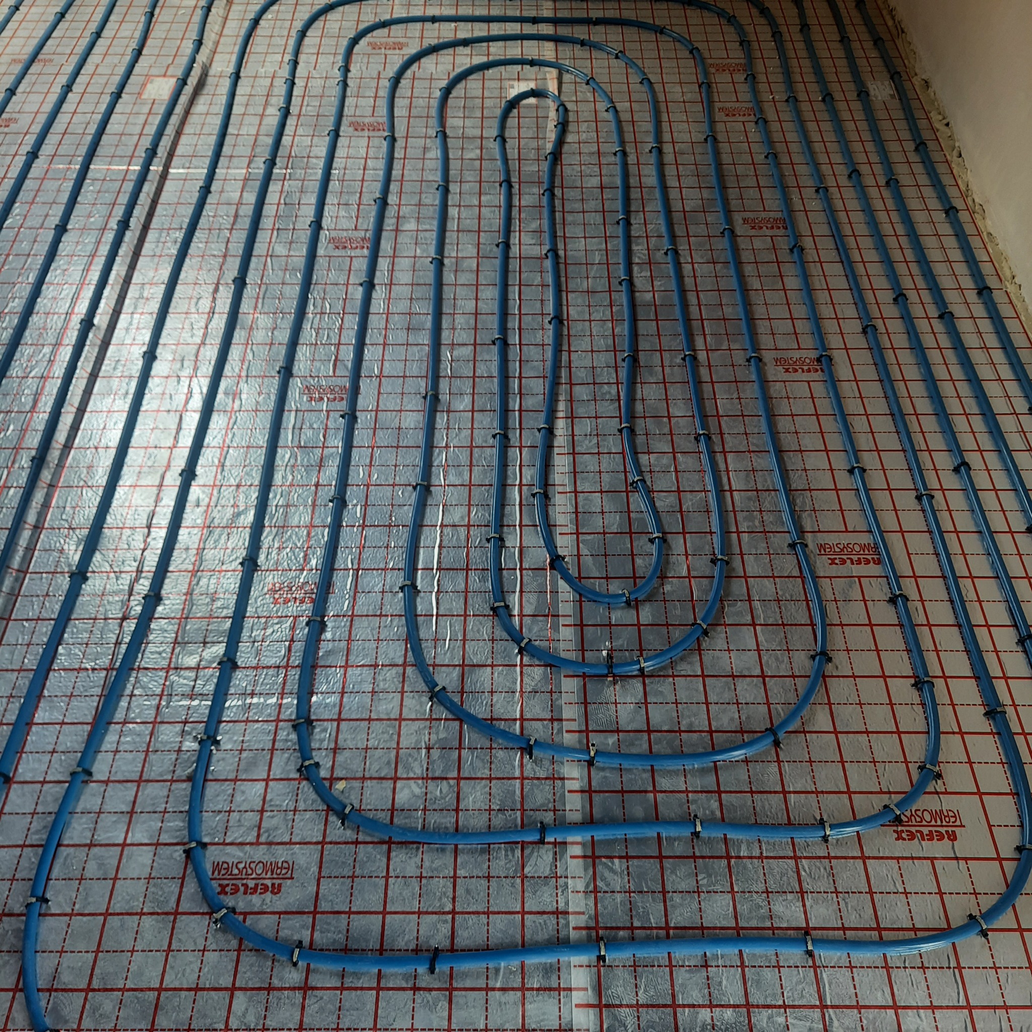 Niebieskie rury ogrzewania podłogowego, przymocowane czarnymi klipsami do srebrnej folii refleksyjnej z czerwonym nadrukiem kratki na podłożu, widok z góry.