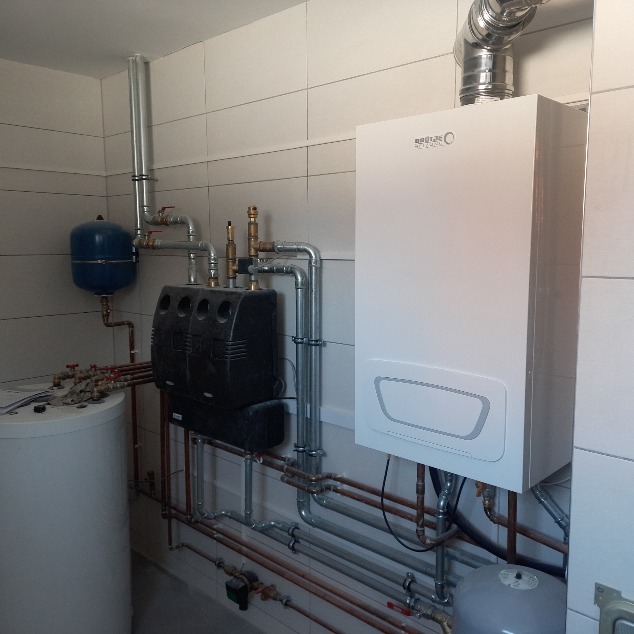 Nowoczesny, biały piec gazowy marki Brötje z podłączeniami hydraulicznymi i gazowymi, widoczne rury miedziane i stalowe, zbiornik wyrównawczy i zasobnik ciepłej wody w pomieszczeniu z białymi...