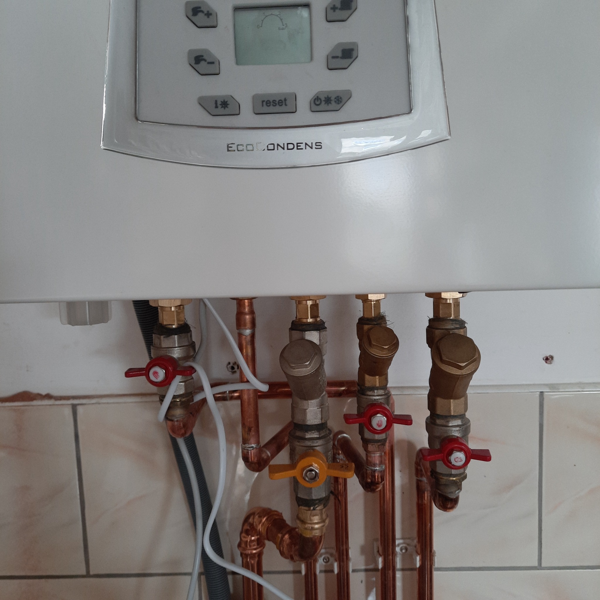 Biały piec gazowy EcoCondens z widocznymi miedzianymi rurami, zaworami z czerwonymi i żółtymi pokrętłami oraz szarym kablem w tle na tle jasnych płytek ceramicznych.