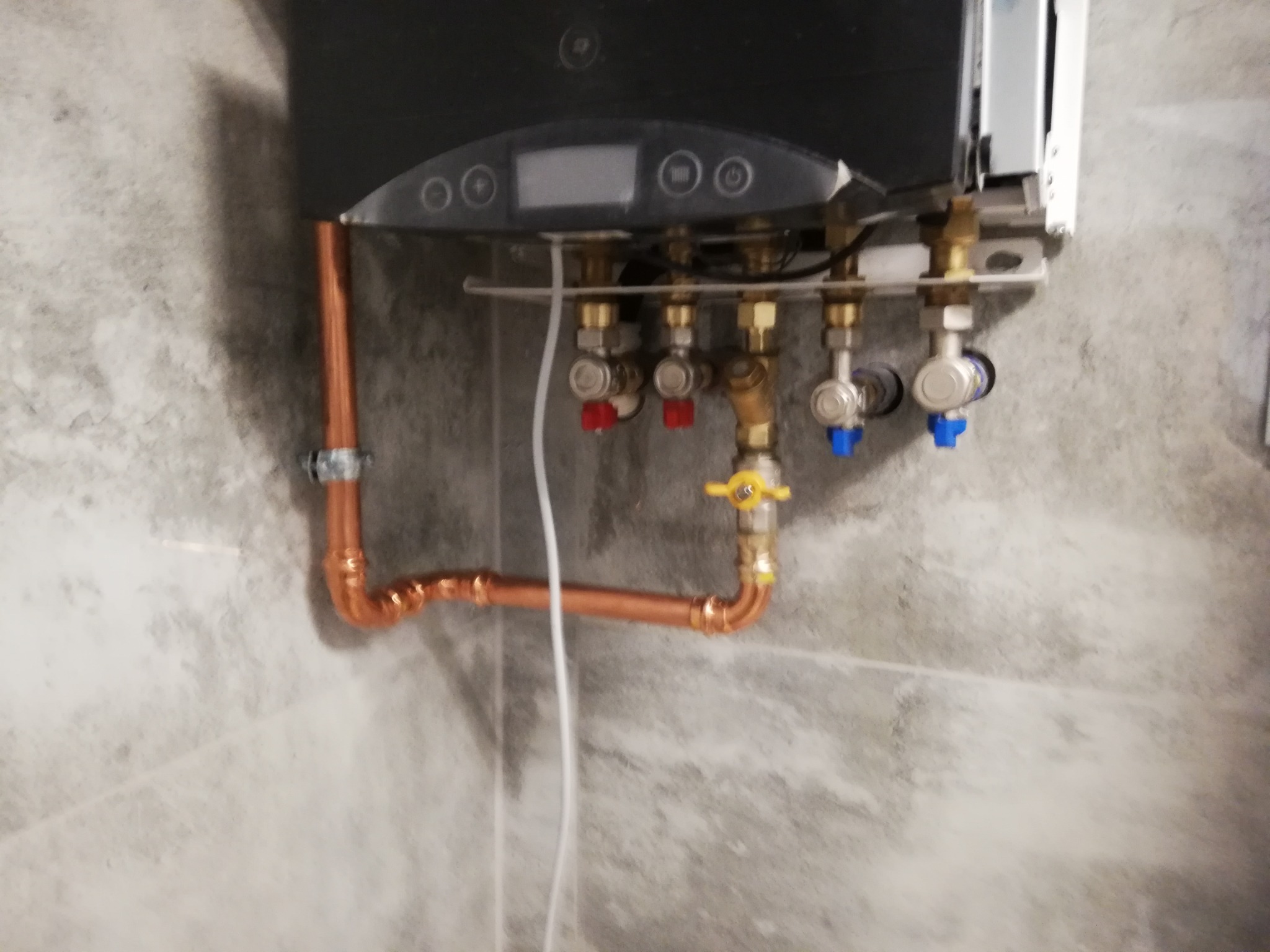 Zamontowany, nowoczesny piec gazowy z miedzianymi rurami i zaworami z czerwonymi i niebieskimi pokrętłami na tle ściany z szarego marmuru.