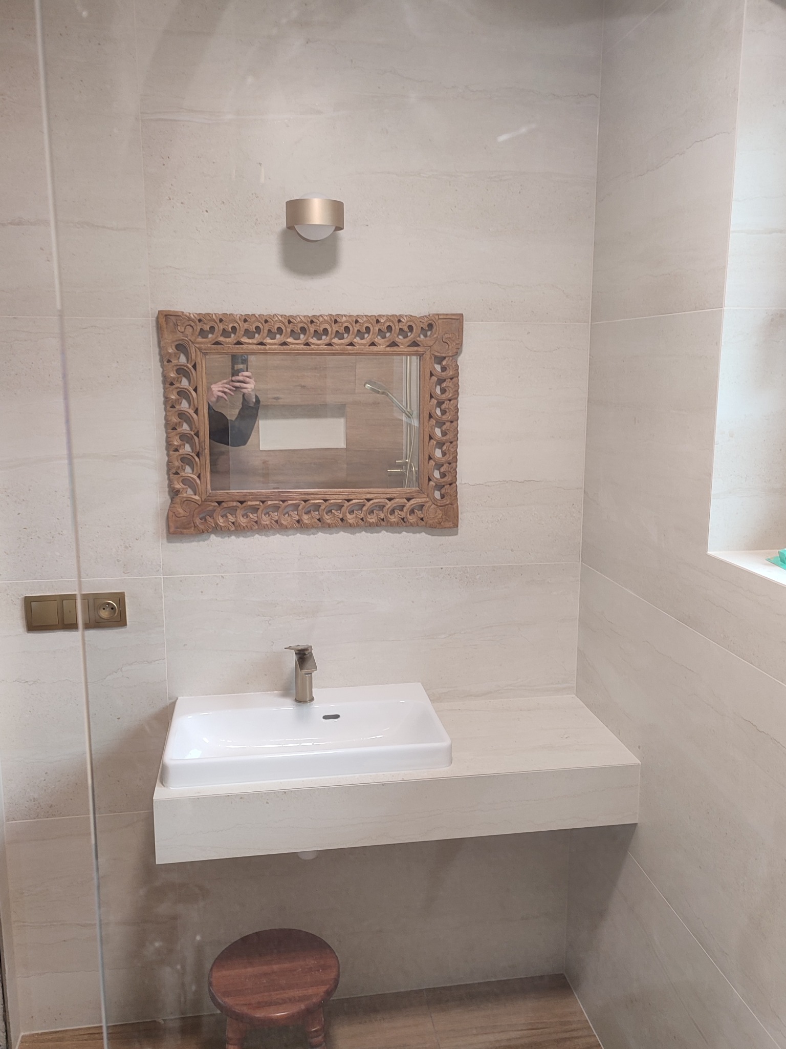 Wykończona łazienka w beżowych płytkach z umywalką na blacie, ozdobnym lustrem w drewnianej ramie i małym stołkiem. Widoczne gniazdka i włączniki w kolorze złotym.