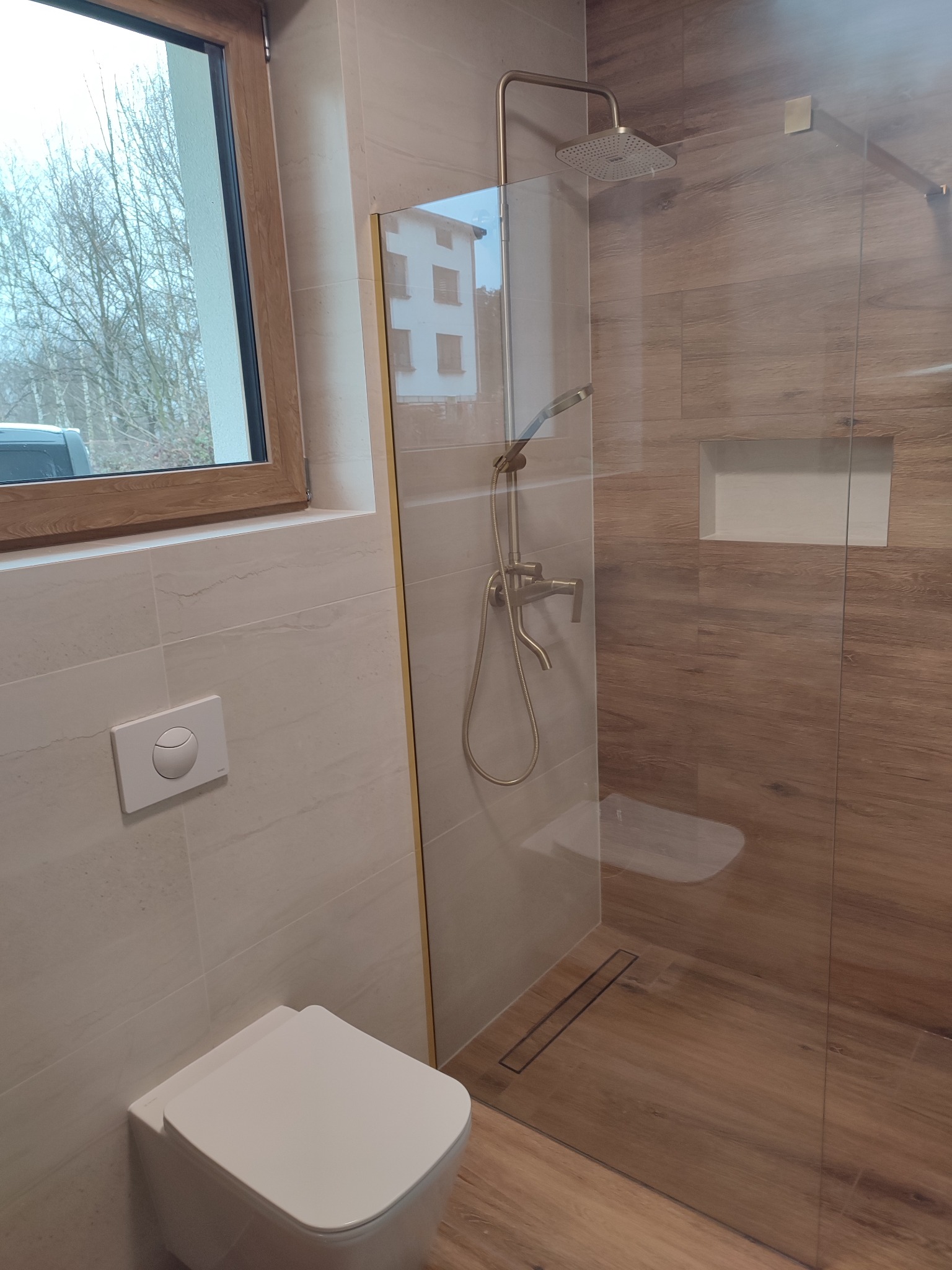 Nowoczesna łazienka z kabiną prysznicową typu walk-in, wykończona płytkami imitującymi drewno i beżowym gresem. Widoczny biały sedes podwieszany i okno.