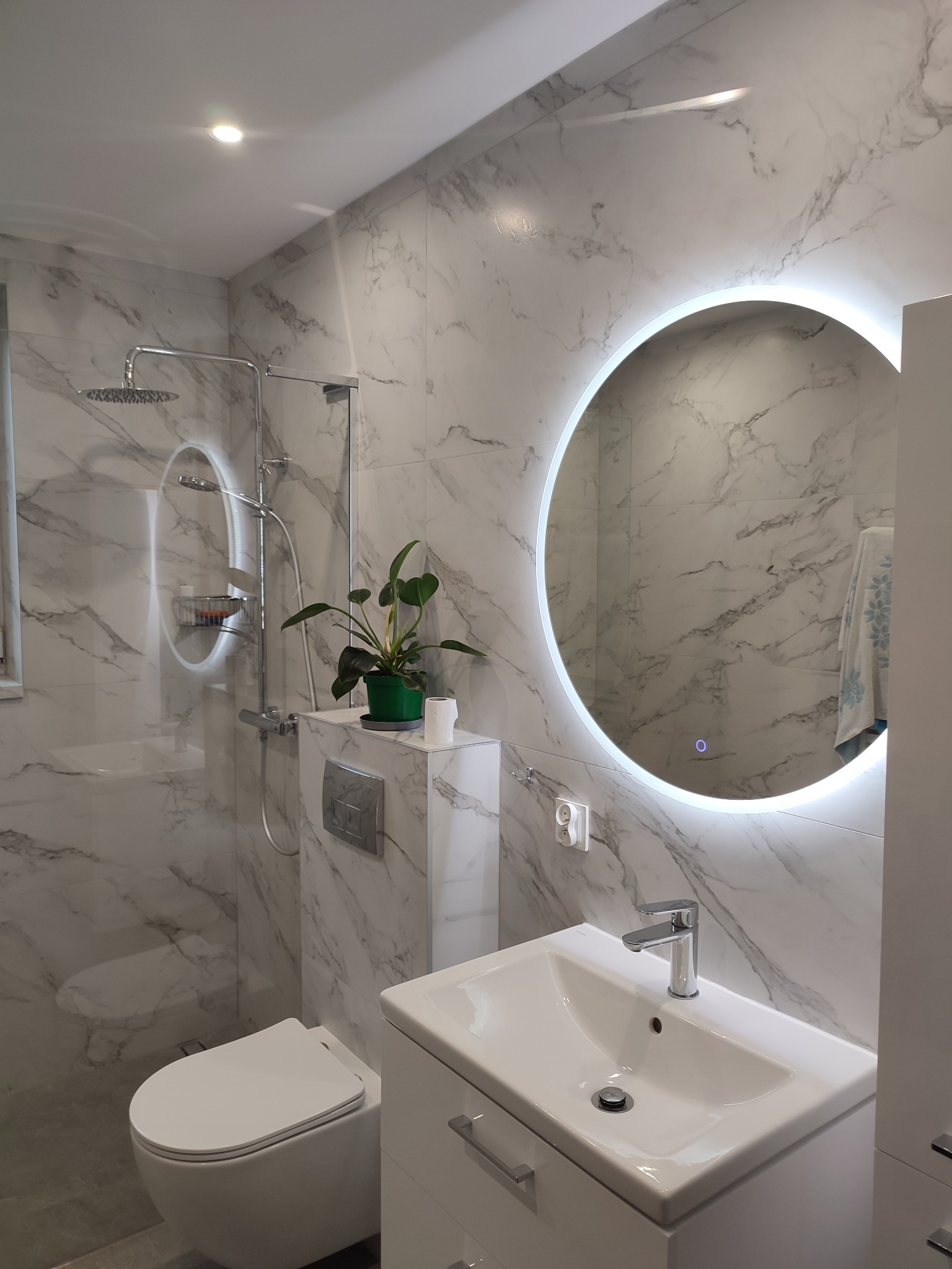 Nowoczesna łazienka z marmurowymi płytkami, okrągłym lustrem z podświetleniem LED, białą umywalką z szafką i toaletą. Minimalistyczny design z akcentem roślinnym.