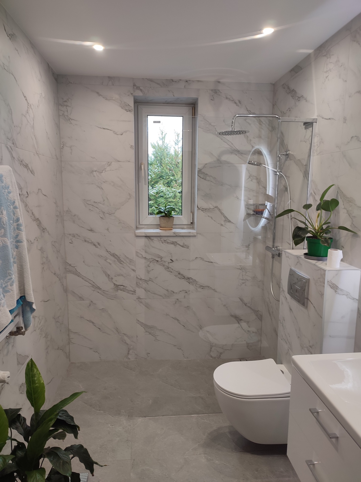 Nowoczesna łazienka z marmurowymi płytkami, kabiną prysznicową typu walk-in i wiszącą toaletą. Rośliny doniczkowe dodają świeżości wnętrzu. Widok z okna na zieleń.