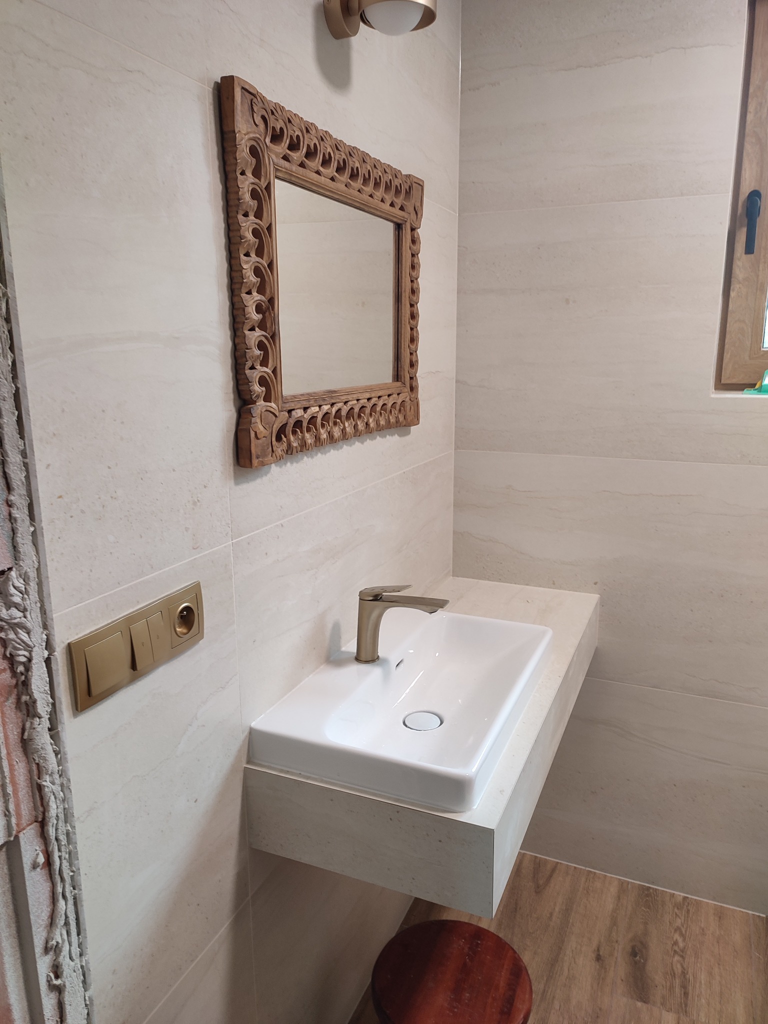 Wykończona łazienka z beżowymi płytkami, ozdobnym lustrem w drewnianej ramie i białą umywalką z baterią w kolorze złota. Widoczne gniazdka i fragment niedokończonej ściany.