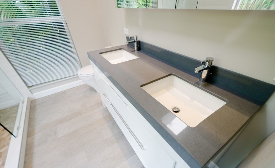 Nowoczesna łazienka z podwójną umywalką nablatową na szarym blacie, białe szafki z chromowanymi uchwytami, jasna podłoga imitująca drewno, okno z żaluzjami w tle.