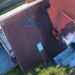 Mycie dachu z czerwonej dachówki z użyciem myjki ciśnieniowej, widoczna różnica między umytą a brudną częścią dachu, ujęcie z drona.