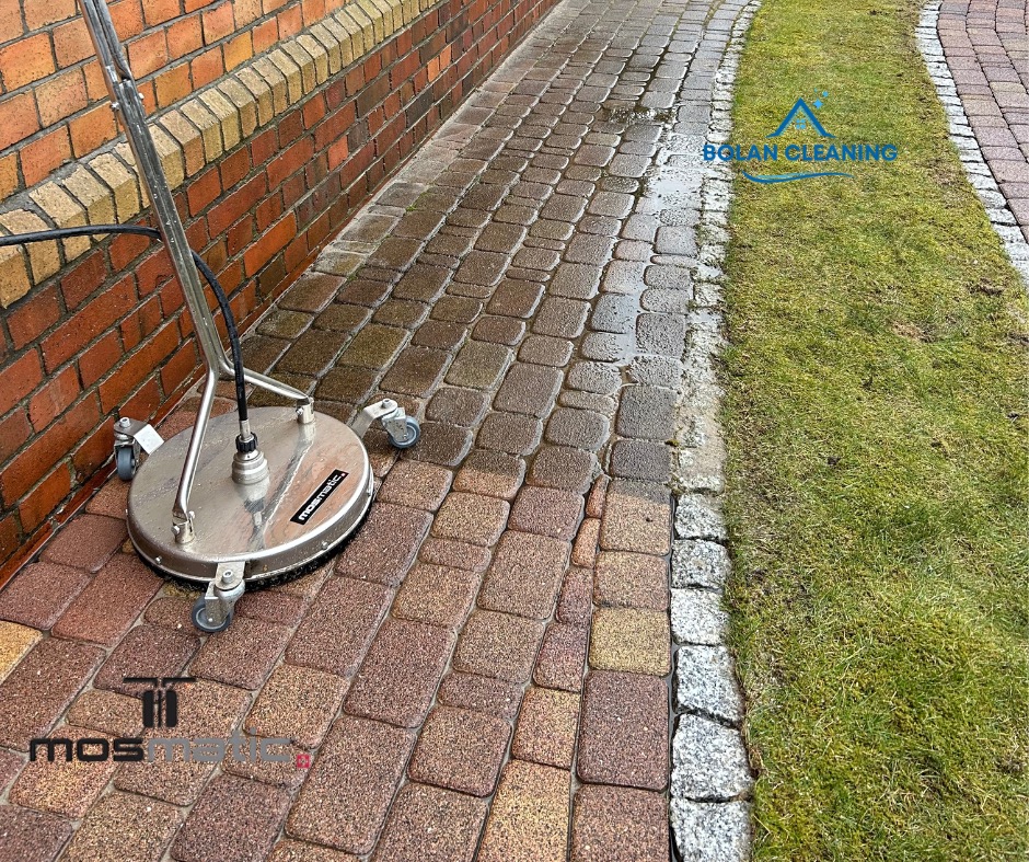 Profesjonalna maszyna do czyszczenia kostki brukowej w trakcie pracy, widoczna mokra i czysta powierzchnia kostki, obok trawnik z obrzeżem.