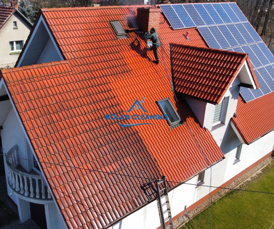 Częściowo wyczyszczony dach z czerwonej dachówki, widoczny pracownik w kombinezonie czyszczący ciśnieniowo okolice komina i okna dachowego, panele słoneczne na dachu, drabina oparta o ścianę budynku.