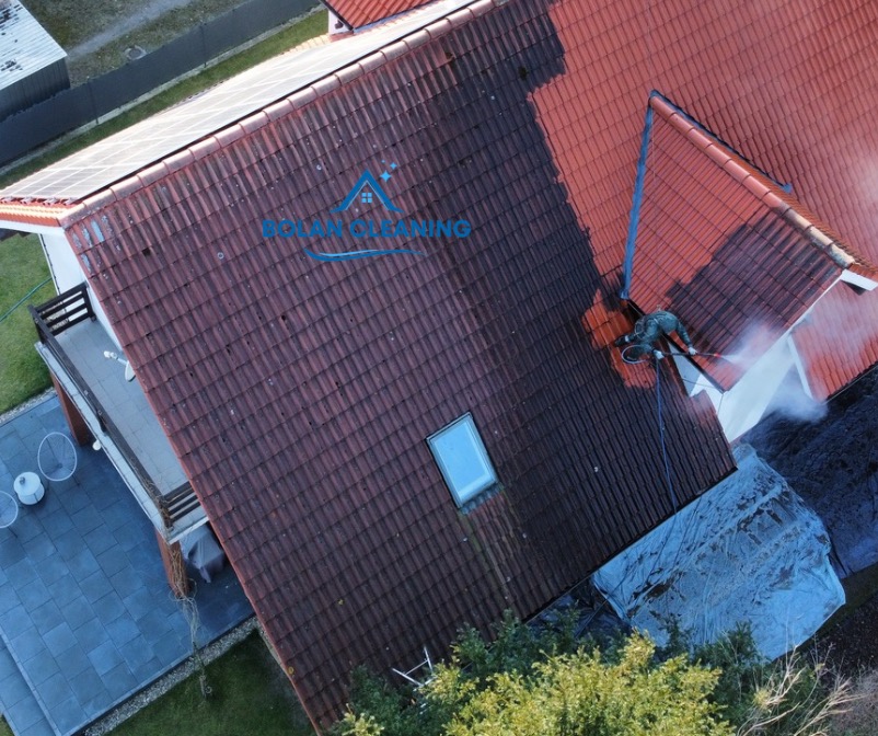 Mycie dachu z czerwonej dachówki z użyciem myjki ciśnieniowej, widoczna różnica między umytą a brudną częścią dachu, ujęcie z drona.