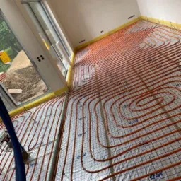 Rozłożona instalacja ogrzewania podłogowego z rur w kolorze pomarańczowym na folii z siatką w jasnym pomieszczeniu z dużym oknem.