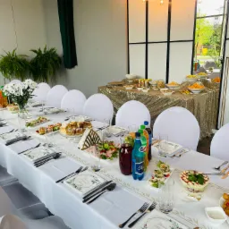 Uroczysty, długi stół z białym obrusem i dekoracjami kwiatowymi, nakryty dla wielu gości, z widocznymi potrawami, napojami i zastawą, w tle bufet ze złotym obrusem i przekąskami.