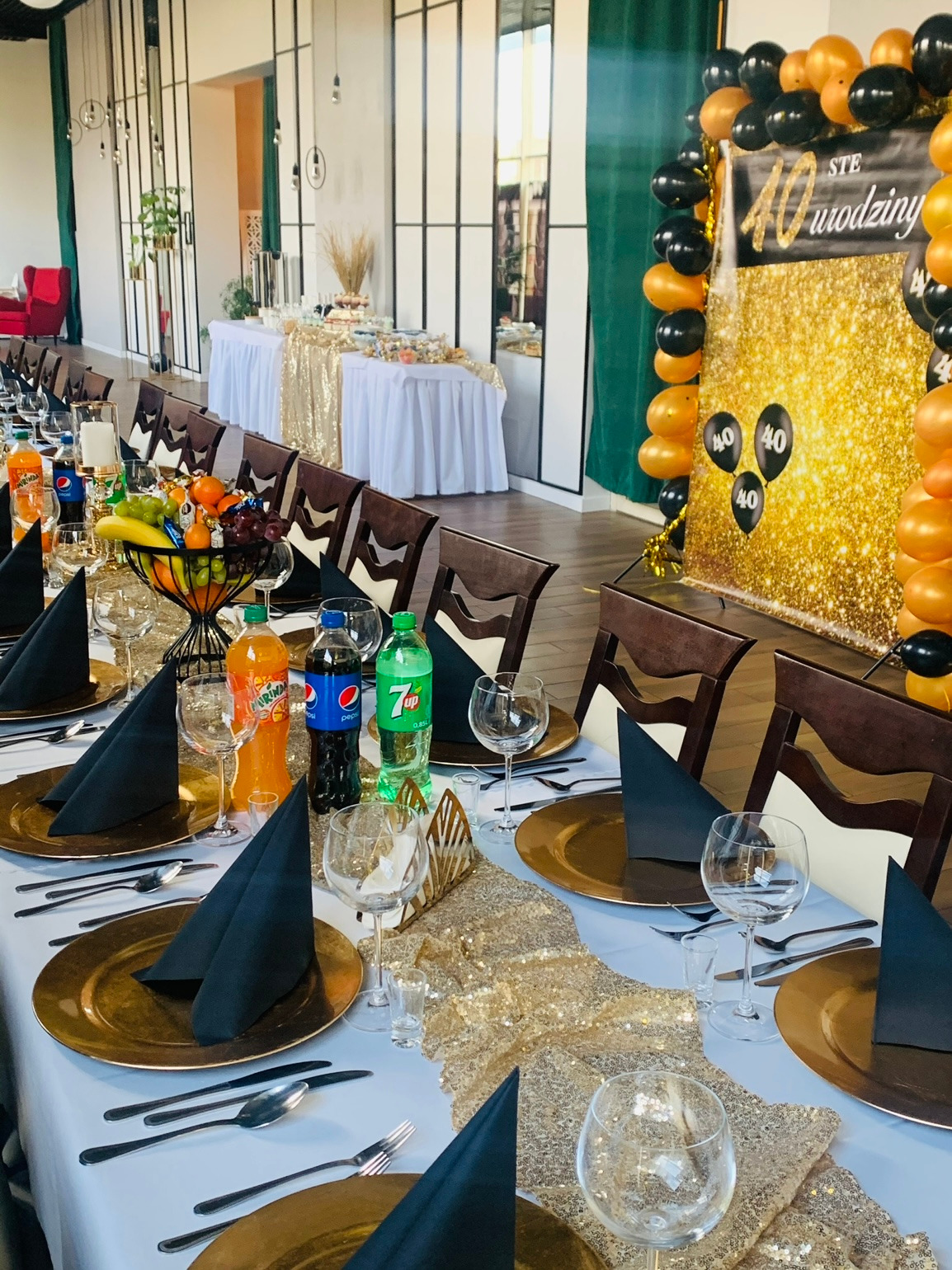 Uroczysty stół na 40 urodziny z złotymi talerzami, czarnymi serwetkami, owocami, napojami, dekoracjami balonowymi i błyszczącym tłem.
