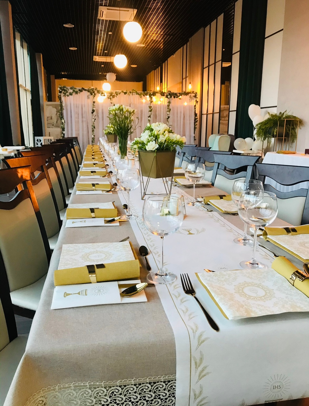 Długi stół bankietowy nakryty dla gości, z białym bieżnikiem i dekoracją kwiatową, przygotowany na uroczystość w przestronnej sali z oknami.