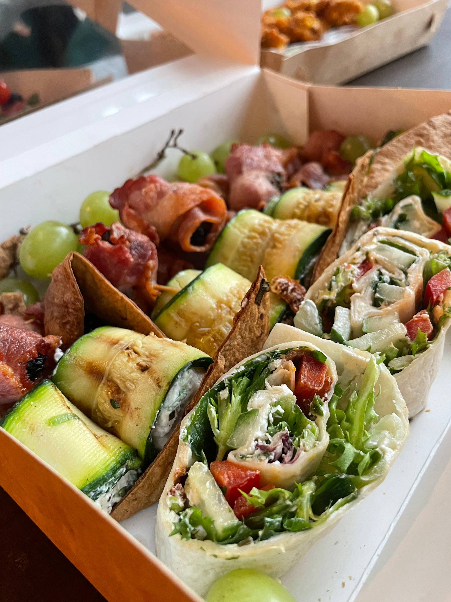 Elegancko zapakowany zestaw przekąsek w papierowym pudełku: tortille z warzywami, roladki z cukinii z białym serem oraz szaszłyki z winogron i bekonu.