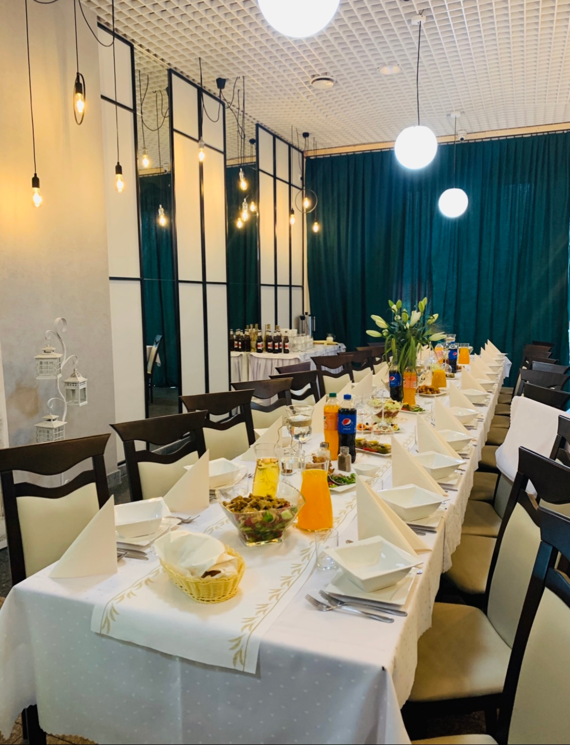 Uroczysty stół bankietowy z białym obrusem i zastawą, przygotowany na imprezę w Radomiu. Widoczne szklane naczynia, napoje w butelkach, sałatki w misach i dekoracje kwiatowe. W tle ciemnozielone...