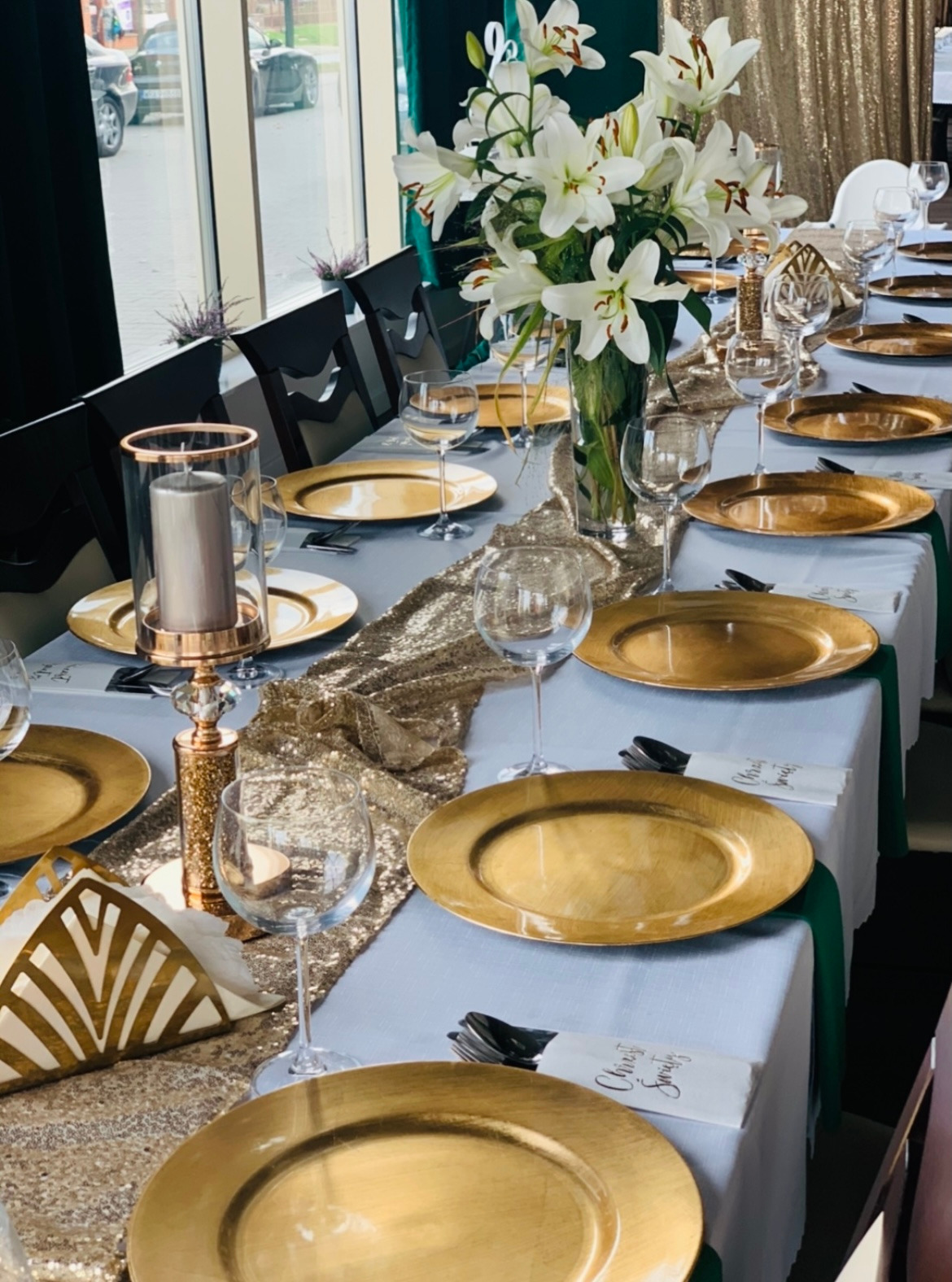 Elegancko nakryty stół z złotymi talerzami, białymi liliami w wazonie, świecami w szklanych lampionach i złotym bieżnikiem na jasnoniebieskim obrusie. Ustawione kieliszki do wina i serwetki z napisem.