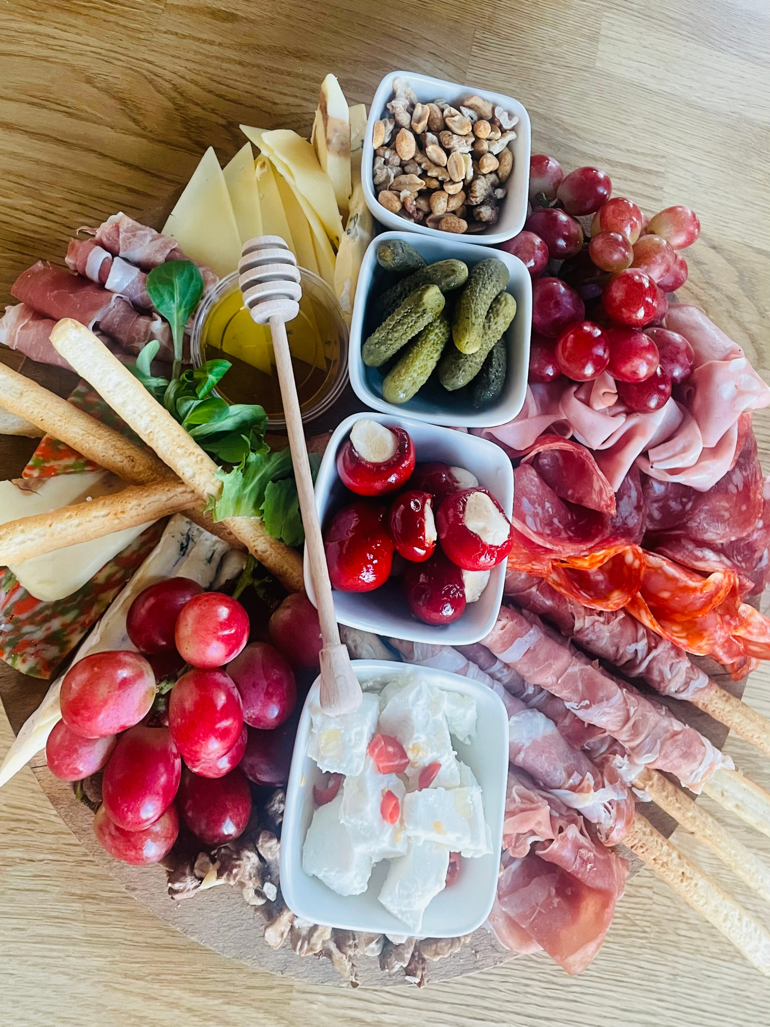 Elegancka deska z przekąskami: sery, szynka parmeńska owinięta wokół paluszków, winogrona, pikle, papryczki faszerowane, mozzarella z pomidorami, orzechy, miód z nabierakiem, na drewnianym blacie.