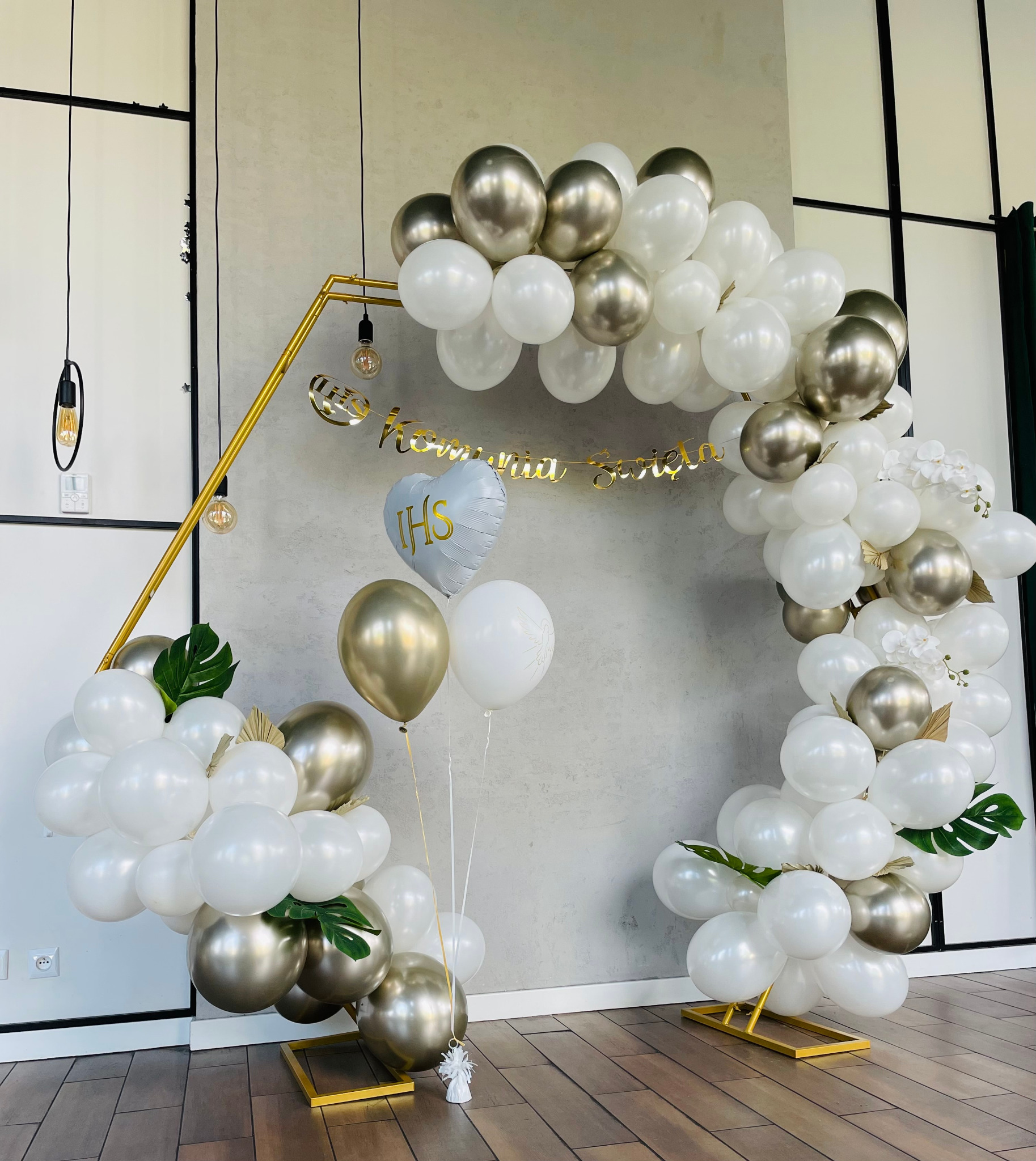 Dekoracja z białych i złotych balonów w kształcie łuku, z napisem 'IHS Komunia Święta' zawieszonym na złotym stelażu, uzupełniona o zielone liście monstery i białe kwiaty.