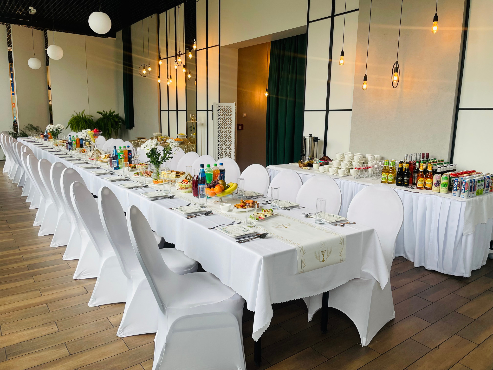 Długi stół bankietowy nakryty białym obrusem z dekoracyjnym bieżnikiem, otoczony krzesłami w białych pokrowcach, zastawiony naczyniami, napojami i przekąskami w jasnym wnętrzu sali z oknami.