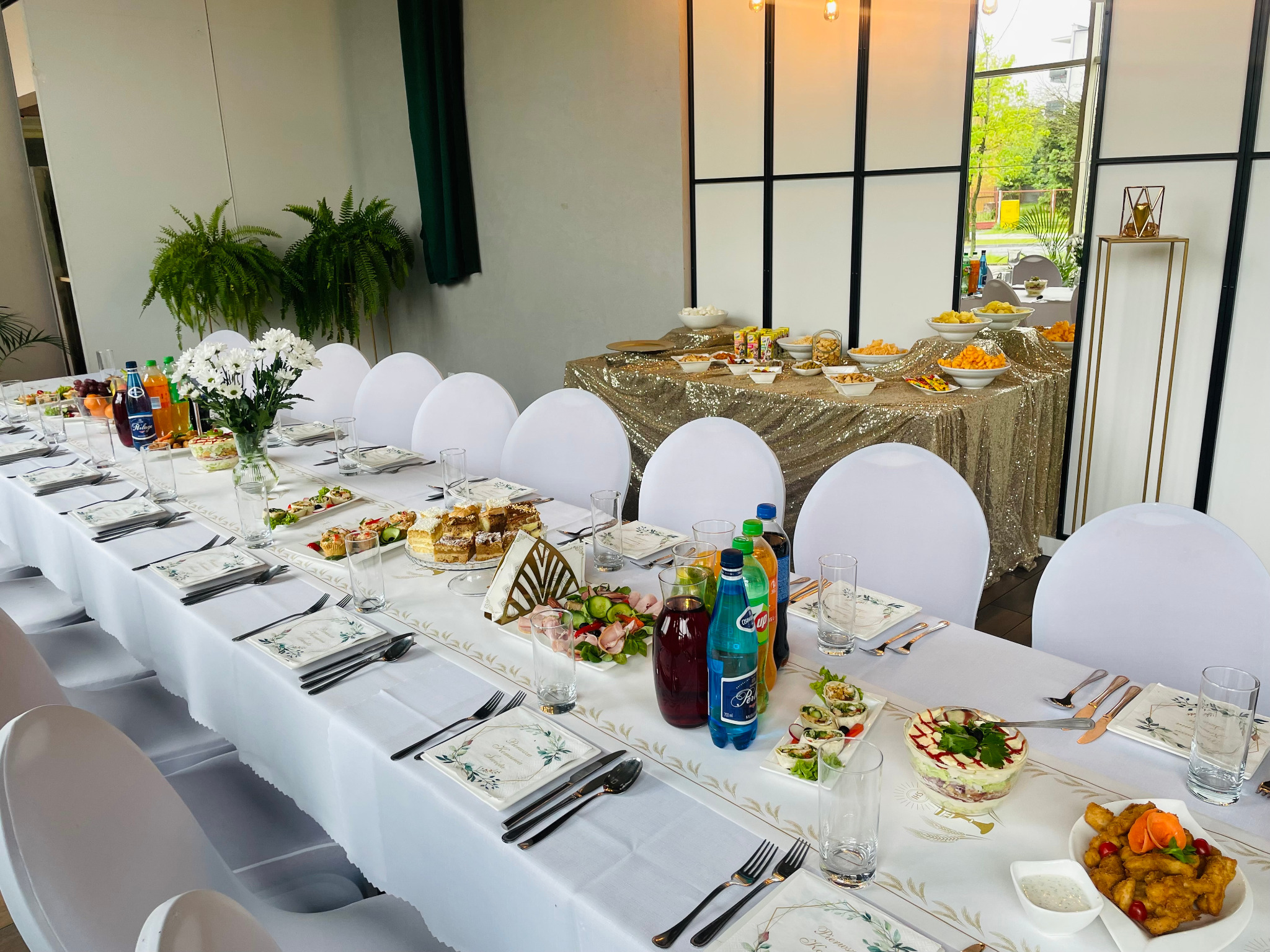Uroczysty, długi stół z białym obrusem i dekoracjami kwiatowymi, nakryty dla wielu gości, z widocznymi potrawami, napojami i zastawą, w tle bufet ze złotym obrusem i przekąskami.