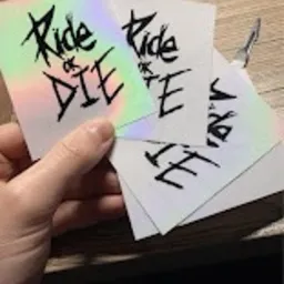 Dłoń trzymająca trzy błyszczące, prostokątne naklejki z napisem 'Ride or Die' w czarnym, grunge'owym stylu. Jedna naklejka ma holograficzne wykończenie, tworzące efekt tęczy.