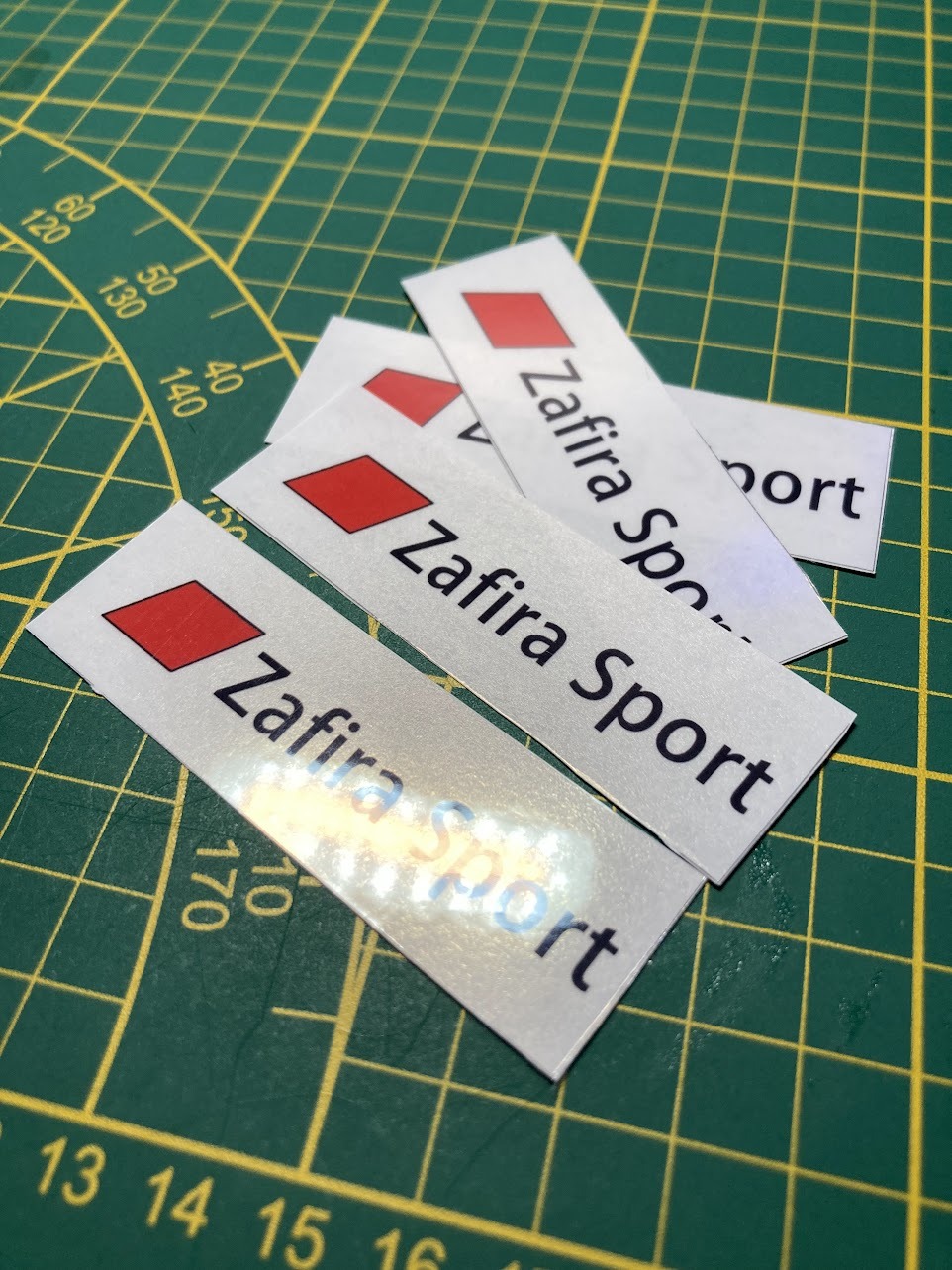 Srebrne naklejki z czerwonym rombem i napisem 'Zafira Sport' ułożone na zielonej macie do cięcia z żółtą siatką.