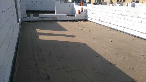 Wnętrze budynku w trakcie budowy z białymi ścianami z betonu komórkowego i przygotowanym podłożem z piasku, widoczne wystające drewniane kołki. Pomarańczowe rury wentylacyjne w tle.