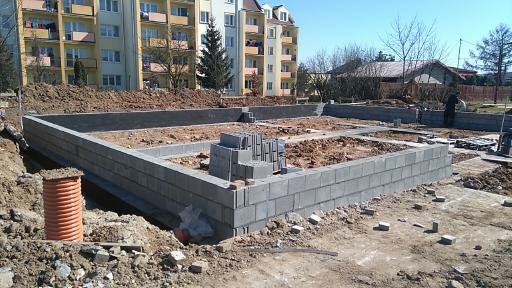 Wznoszenie fundamentów z bloczków betonowych na placu budowy, widoczny pomarańczowy karbowany przewód, w tle budynek mieszkalny i drzewa.