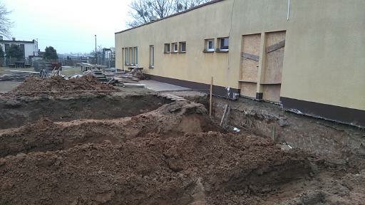 Żółta elewacja budynku w trakcie prac ziemnych; wykopane fundamenty, zabezpieczone deski przy ścianie.