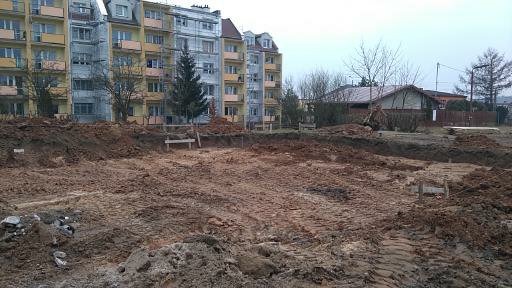 Wykarczowany teren przygotowany pod budowę, widoczne ślady opon i wykopane fundamenty, w tle bloki mieszkalne i dom jednorodzinny.