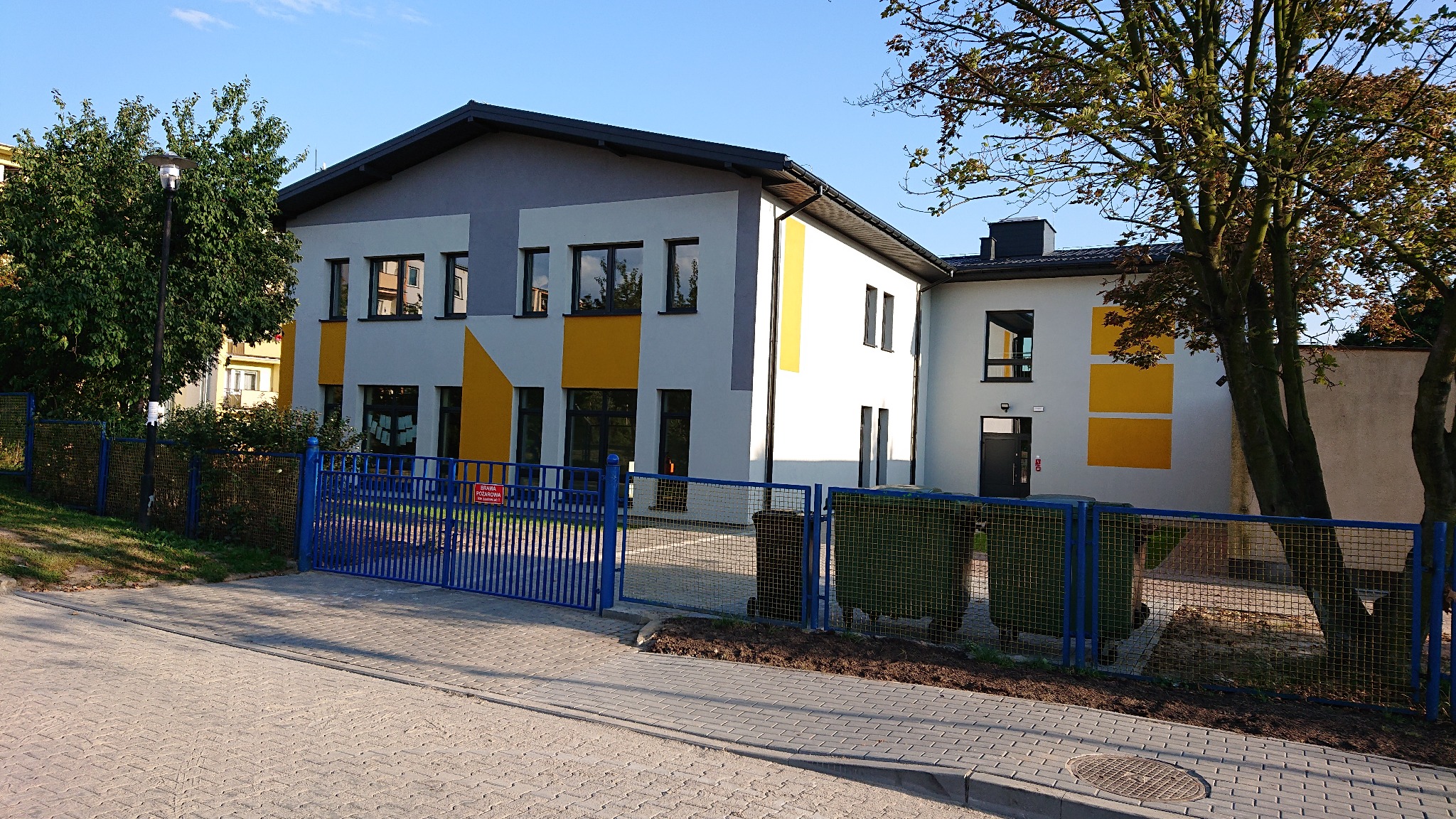 Dwukondygnacyjny budynek pomalowany w szaro-biało-żółte geometryczne wzory, widziany zza niebieskiego ogrodzenia z bramą.