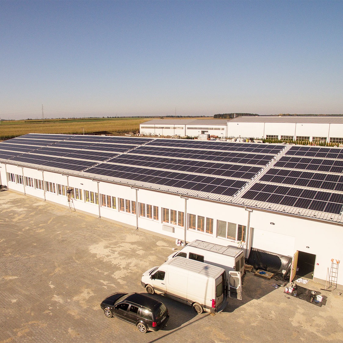 Duża hala z panelami słonecznymi na dachu, widok z góry, z zaparkowanymi samochodami dostawczymi i osobowymi.