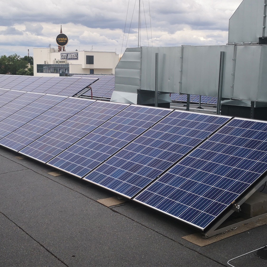 Rząd paneli słonecznych na płaskim dachu budynku, z widocznymi elementami wentylacji i budynkiem hotelowym w tle pod pochmurnym niebem.