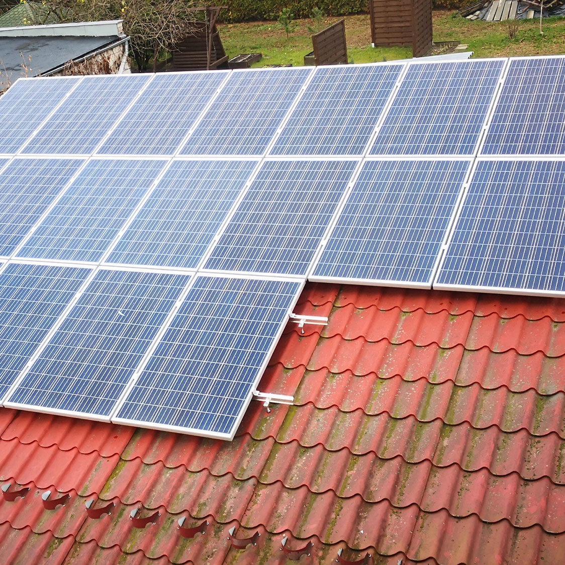 Panele słoneczne częściowo zamontowane na czerwonym dachu pokrytym dachówką ceramiczną, widoczne białe uchwyty montażowe.