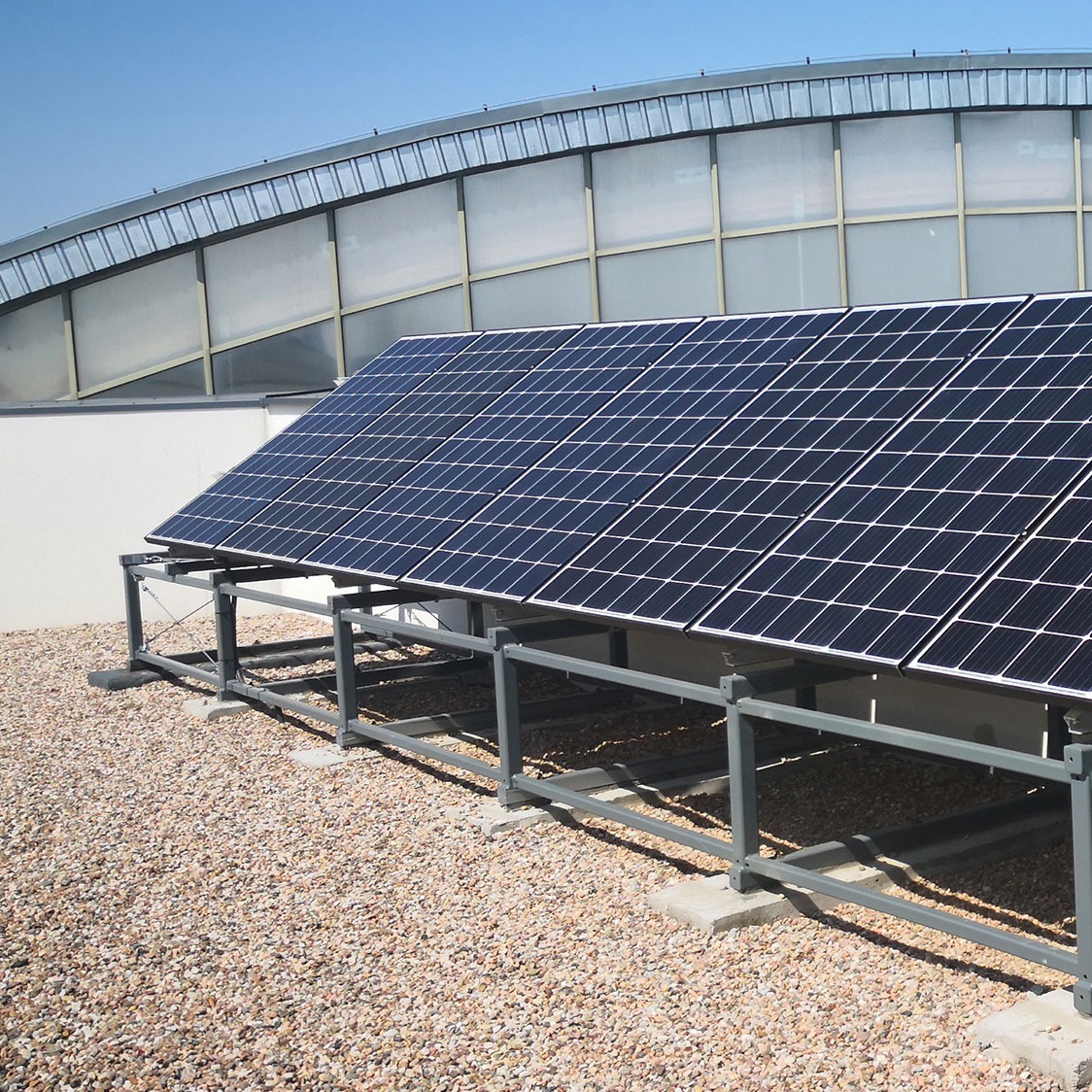 Panele słoneczne na płaskim dachu z metalową konstrukcją wsporczą i żwirowym podłożem, w tle budynek z zaokrąglonym dachem.