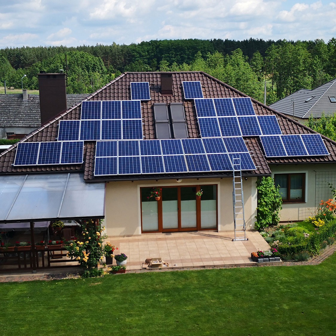 Instalacja fotowoltaiczna na dachu domu jednorodzinnego z brązową dachówką, widoczna aluminiowa drabina oparta o ścianę, zadbany ogród z kwiatami i trawnikiem.